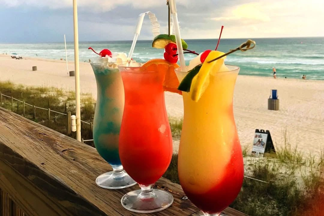 Colorful cocktails sit on a wooden railing with a scenic beach and ocean backdrop at Barefoot Restaurants.