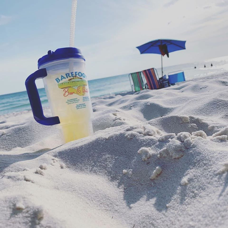 A Barefoot Restaurants cup sits in the soft sand, with a blue umbrella and Gulf waves in the background.